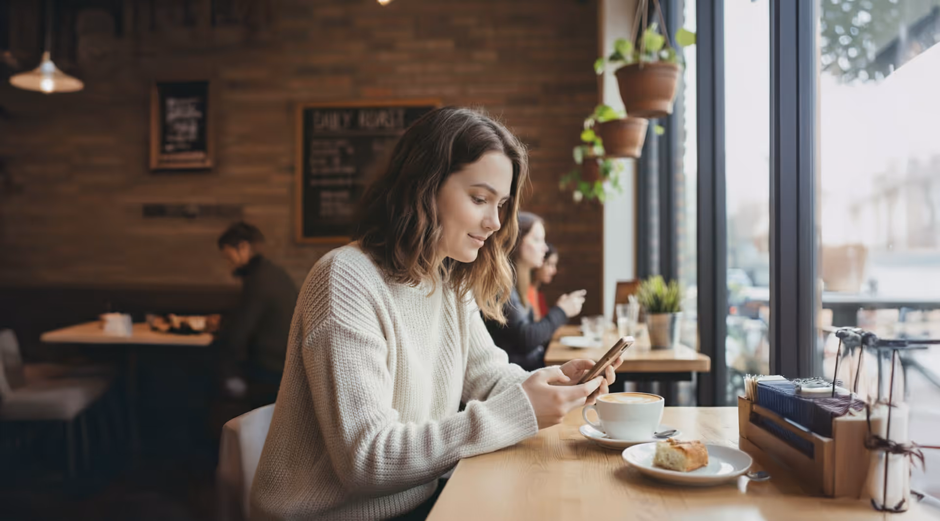 Young woman in a white sweater sitting at a café table by the window, looking at her smartphone with a cup of coffee and a pastry on the table.